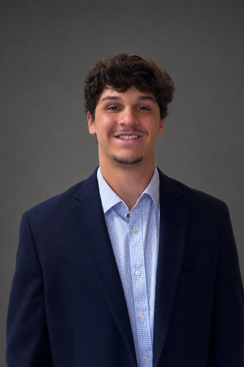 Professional headshot of a smiling man with dark wavy hair wearing a navy blazer and light blue checkered shirt against a gray background